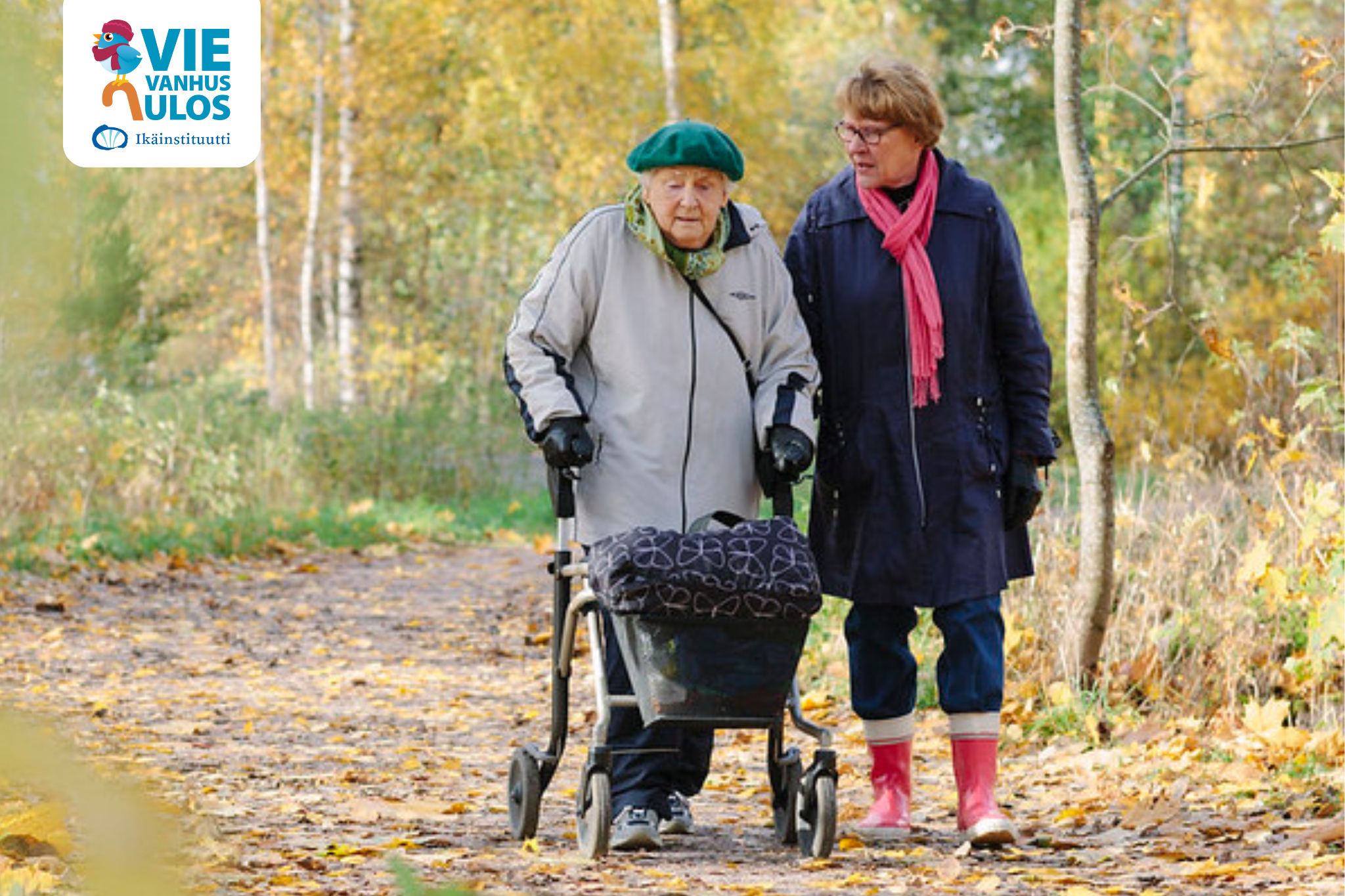Keski-ikäinen nainen ulkoilee iäkkään kanssa. Iäkkäällä on tukenaan rollaattori. Kävelevät syksyisessä metsässä.