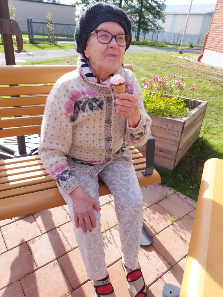 Iäkäs nainen istuu ulkona penkillä ja syö jäätelöä. Naisella silmälasit ja myssy päässä, vieressä kukkalaatikko.