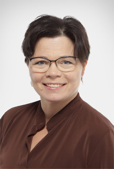 Merja, tummat lyhyet hiukset, silmälasit, katsoo kameraan ja hymyilee. Ruskea paita.