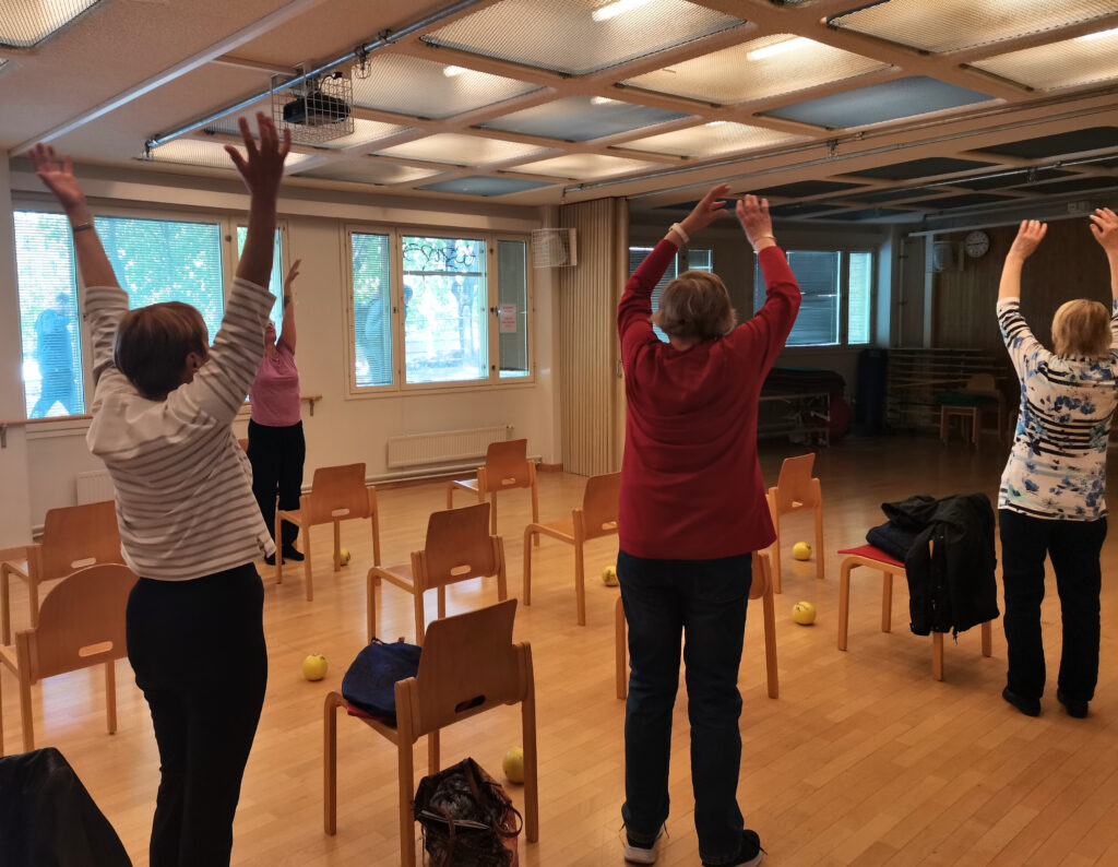 Ihmisiä jumppaamassa salissa, kädet ylhäällä, ohjaaja edessä. Kuvattu ryhmää takaapäin. Lattialla myös tuoleja.