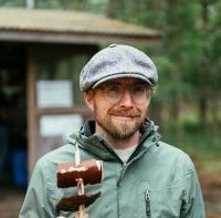 Miikka vuorinen, lakki päässä, silmälasit, partaa, kädessä kaarnalaiva. 