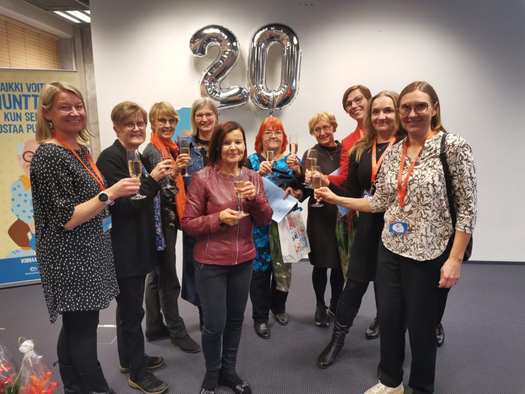 10 ihmistä rivissä, käsissä juhlamalja, katseet kameraan. Taustalla 20-vuotisilmapallot.