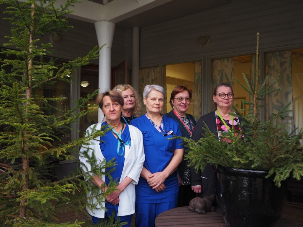 Viisi naista seisoo kahden kuusen välissä. Osalla heistä on hoitajan työvaatteet päällä.