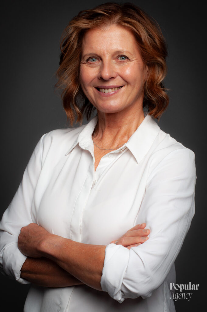 Jaana, ruskeat puolipitkät hiukset, valkoinen paita, katsoo kameraan hymyillen.