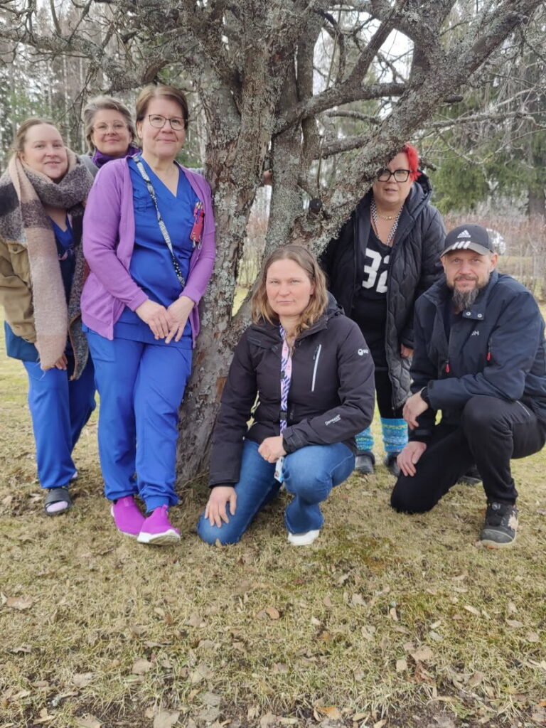 Projektiryhmä puun juurella. Osalla on hoitajan vaatteet ja osalla omat.