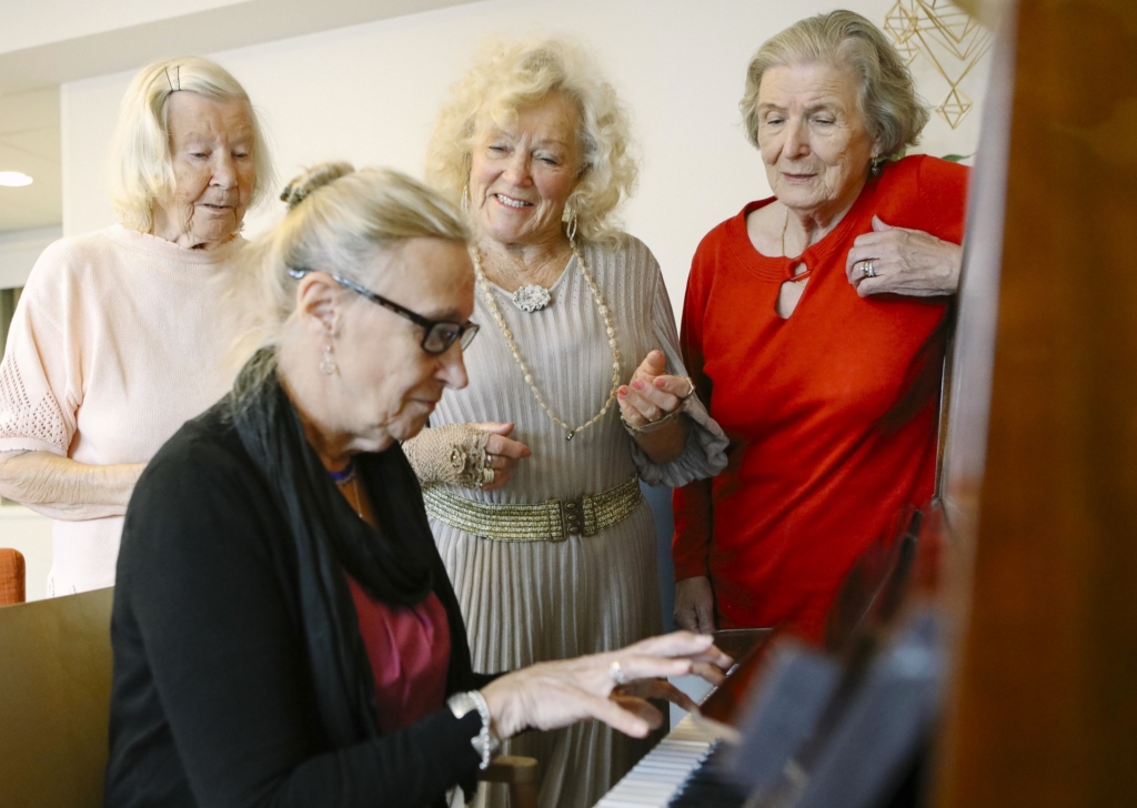 Iäkäs nainen soittaa pianoa ja muut naiset nauttivat musiikista ympärillä.