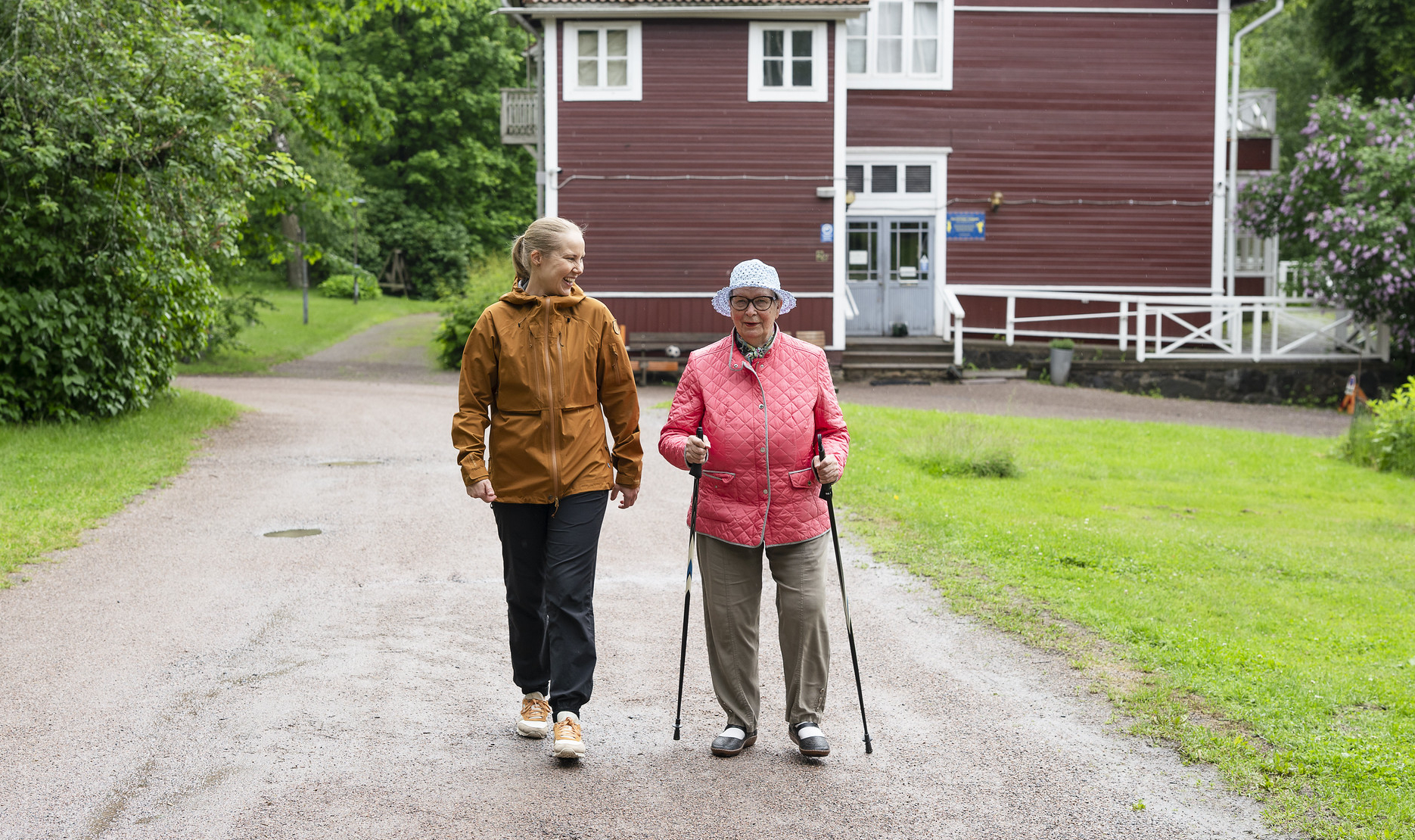 iäkäs ja nuori nainen kävelemässä, iäkkäällä kävelysauvat Taustalla puita, punainen rakennus ja nurmea.