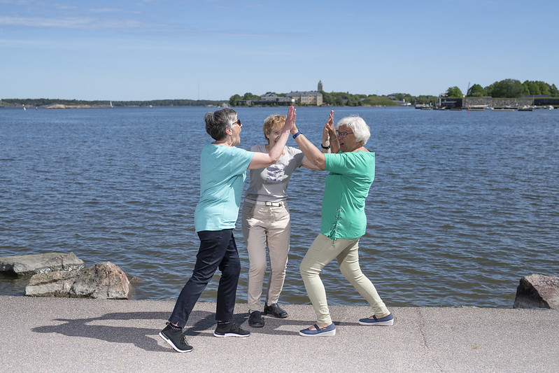 Kolme iäkästä henkilöä rannalla lyövät käsiä yhteen. Taustalla merta ja taivasta.